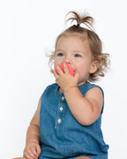 A toddler girl with curly light brown hair in a high ponytail, wearing a blue denim-look sleeveless top, sitting against a plain white background while biting a bright red teething toy.