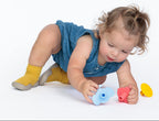 A toddler in a blue denim romper and yellow socks crawls on a white studio floor, playing with silicone teether toys.