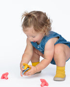A toddler girl in a blue denim dress and yellow socks crouches on a white studio floor, holding a silicone children's teether and silicone monkey Whine Stoppers are scattered around her.