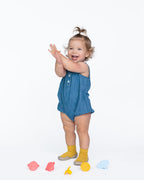 A toddler stands on a white studio background wearing a sleeveless blue denim romper and yellow socks, with small colorful silicone Whine Stoppeers on the floor nearby.