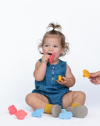 A toddler girl wearing a denim romper and yellow socks sits on a white studio floor playing with colorful silicone teething toys. A hand on the right holds a yellow toy.
