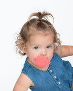 A toddler with blonde hair in a high ponytail wearing a denim top and holding a red monkey-shaped teether, photographed against a plain white background.